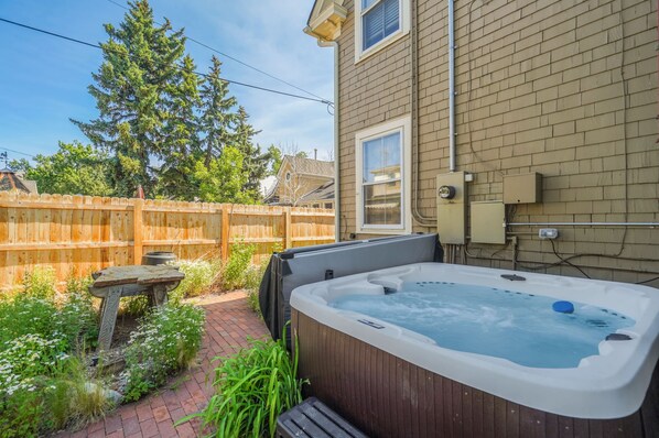 Un jacuzzi en un patio trasero con una cerca de madera, un patio de ladrillo y una casa al fondo.