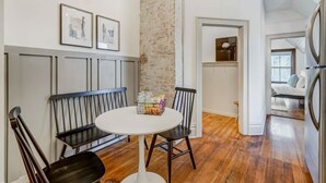 A dining area with a round table, black chairs, and a bench. There are framed pictures on the wall, a brick accent wall, and a view into another room with a bed and a window. - Noble Summit BBQ & Fire Pit  & Scenic Views (Colorado Springs)