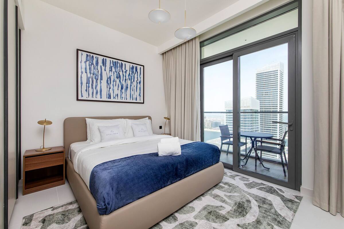 A modern hotel room with a large bed, a view of the city through a sliding glass door, and a cityscape visible through the window.