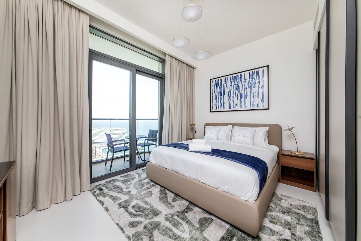 A modern hotel room with a large bed, a view of the city, and a balcony with chairs.