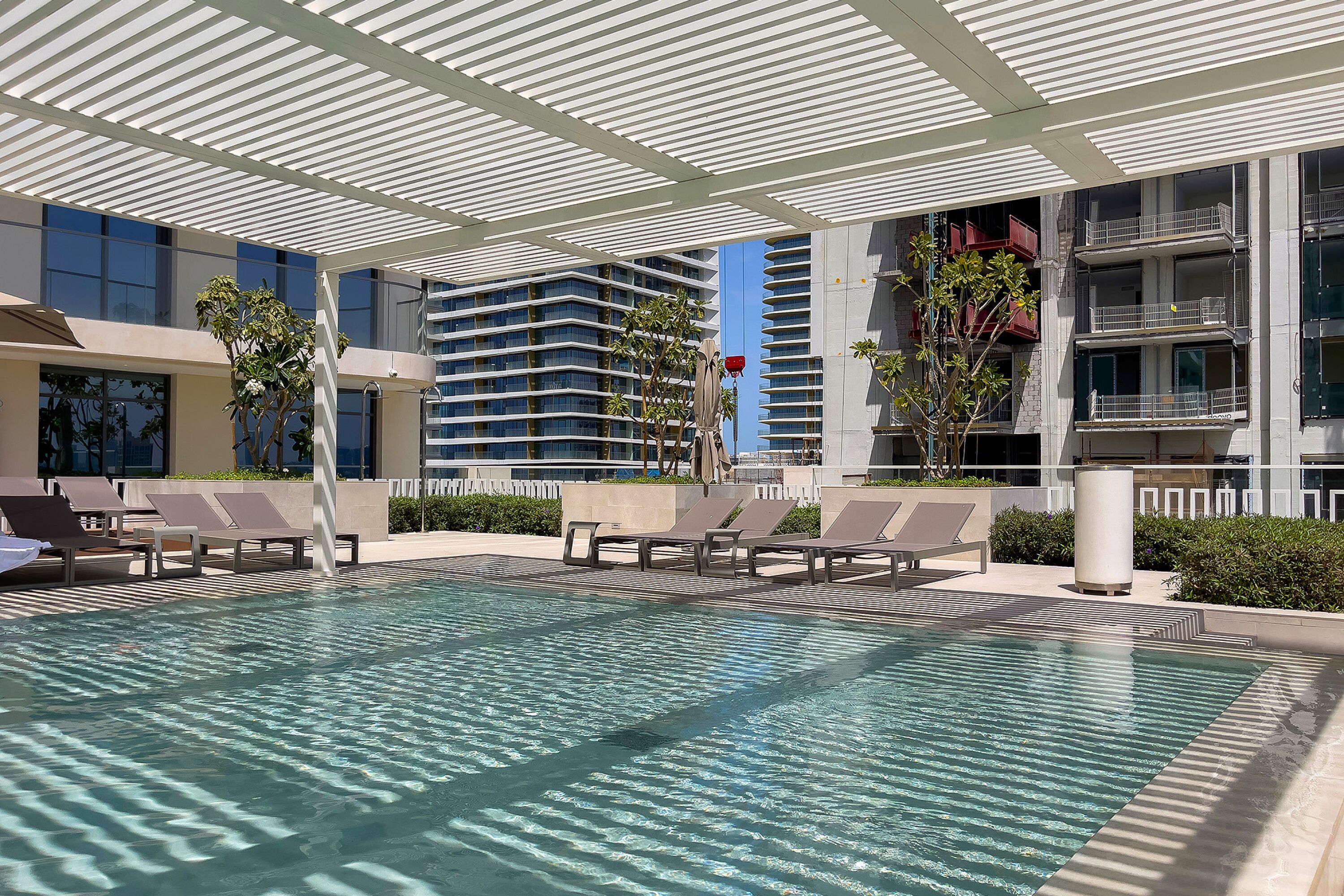 Zona de piscina con sillas de descanso, un área de estar cubierta y un edificio moderno al fondo.