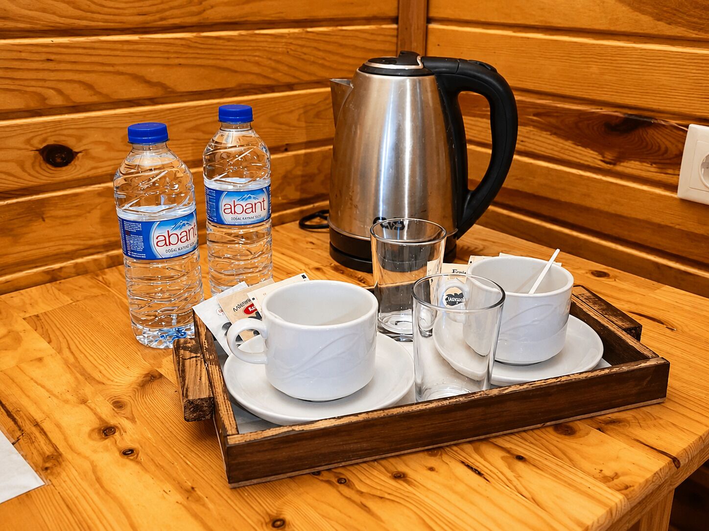 Una mesa de madera con una bandeja que contiene una tetera, dos botellas de agua, dos tazas y dos vasos.