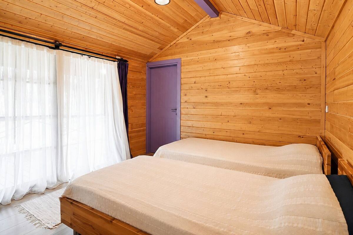 Ein Schlafzimmer einer Holzhütte mit zwei Betten, einem Fenster mit durchsichtigen Vorhängen und einer violetten Tür.