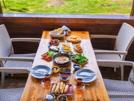 Una mesa de madera dispuesta con diversos platos, incluyendo pan, frutas y un cuenco de aceitunas, en un entorno exterior con sillas de mimbre.