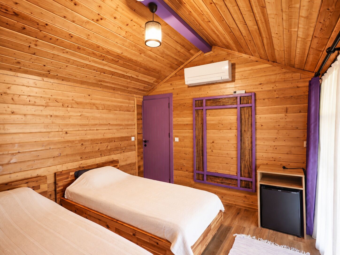 A wooden cabin room with two beds, a purple door, a small black refrigerator, and an air conditioning unit.