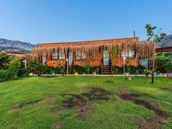 Um edifício de madeira com telhado de palha, rodeado por um jardim bem cuidado com diversas plantas e céu claro.
