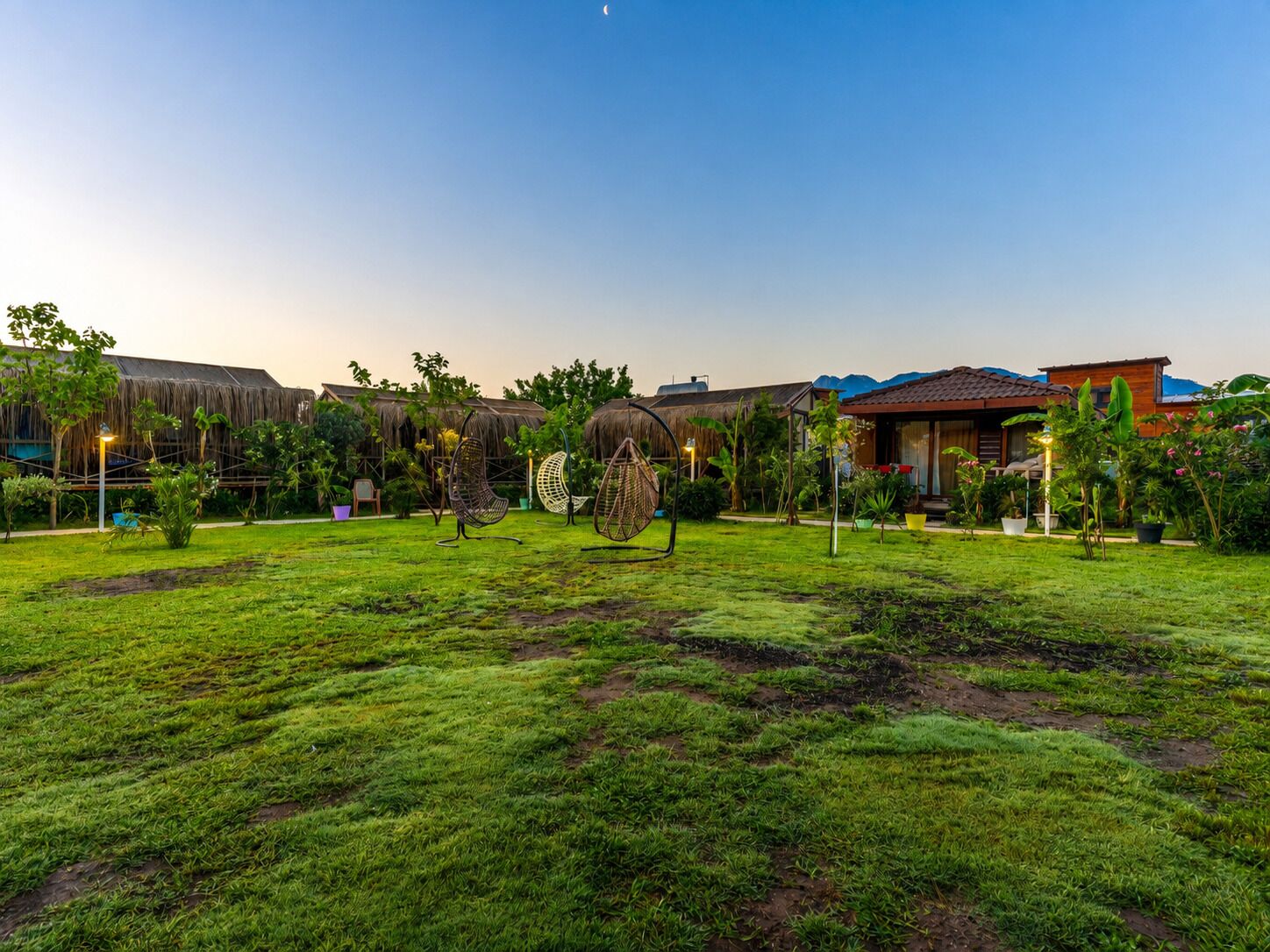 A well-maintained garden with thatched-roof structures, a grassy lawn, and a mountain backdrop.
