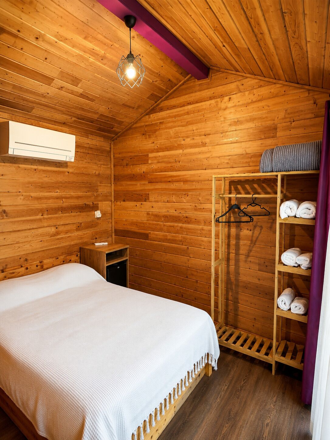Una habitación de madera con una cama, un estante con toallas y una lámpara colgante.