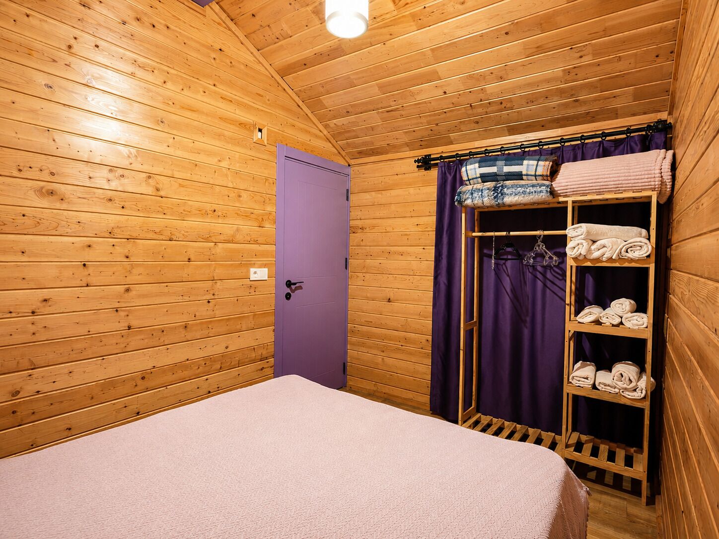 A wooden cabin room with a bed, a purple door, and a shelf with towels.