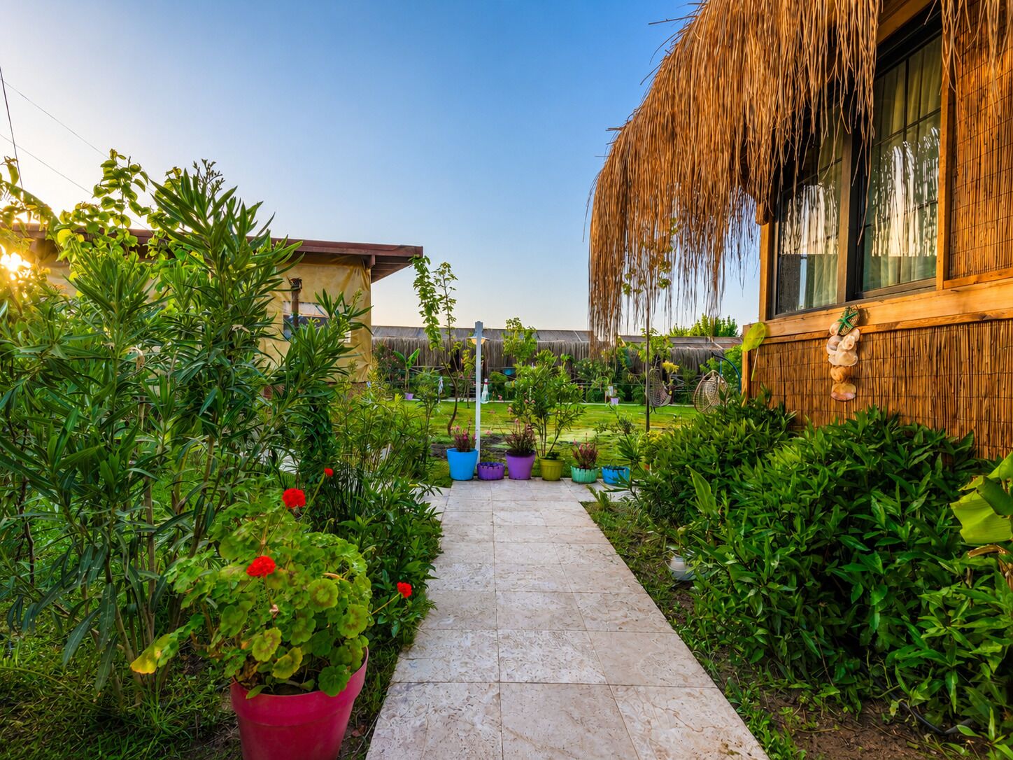Un sendero en un jardín conduce a un edificio con tejado de paja, rodeado de plantas en macetas y vegetación.
