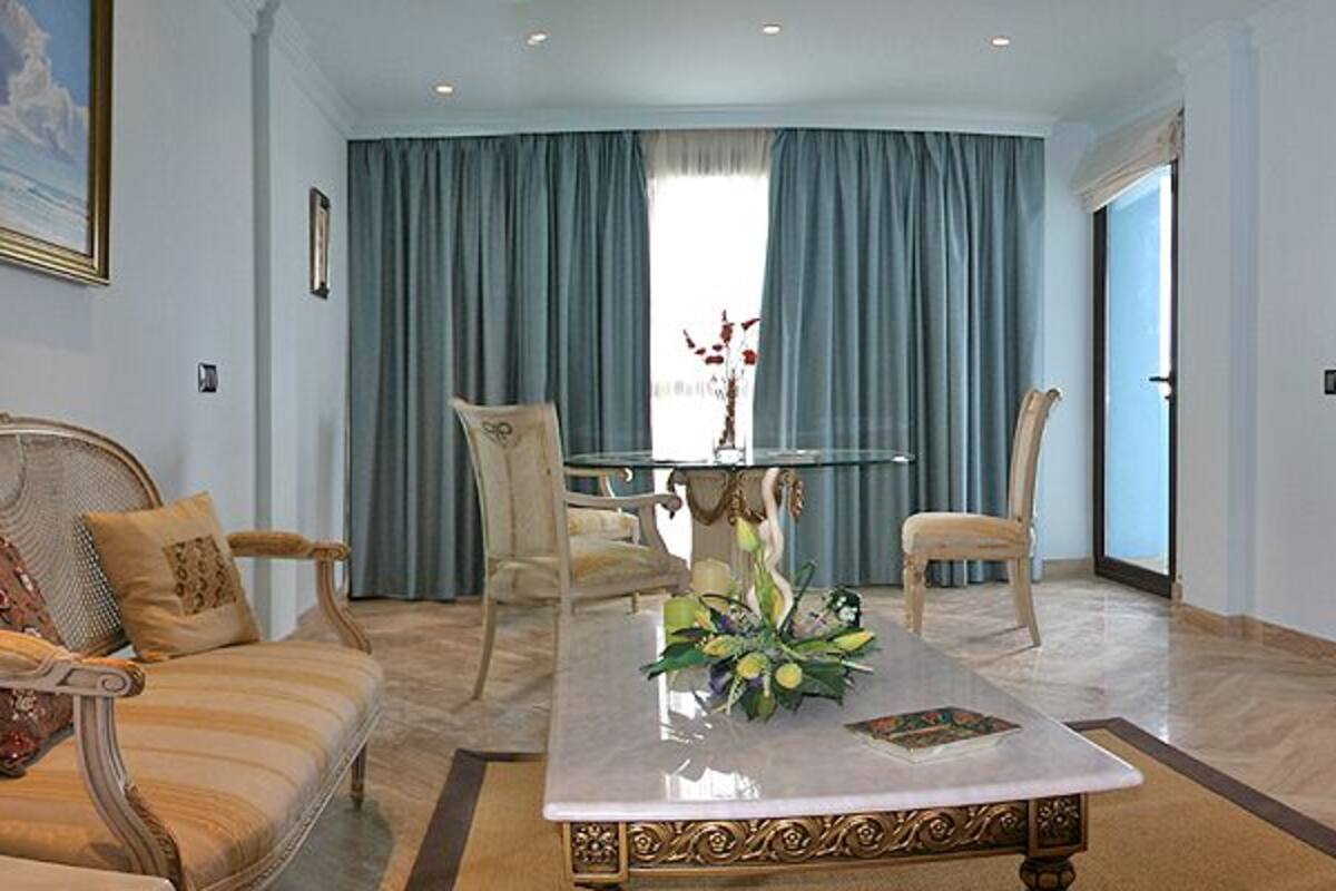 A spacious living room with a sofa, armchair, and a coffee table. There is a television on a cabinet, and a dining table with chairs. The room has a large window with curtains.