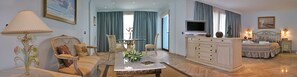 A spacious living room with a sofa, armchair, and a coffee table. There is a television on a cabinet, and a dining table with chairs. The room has a large window with curtains. - Hotel Hacienda Don Manuel  (Granja de Torrehermosa)