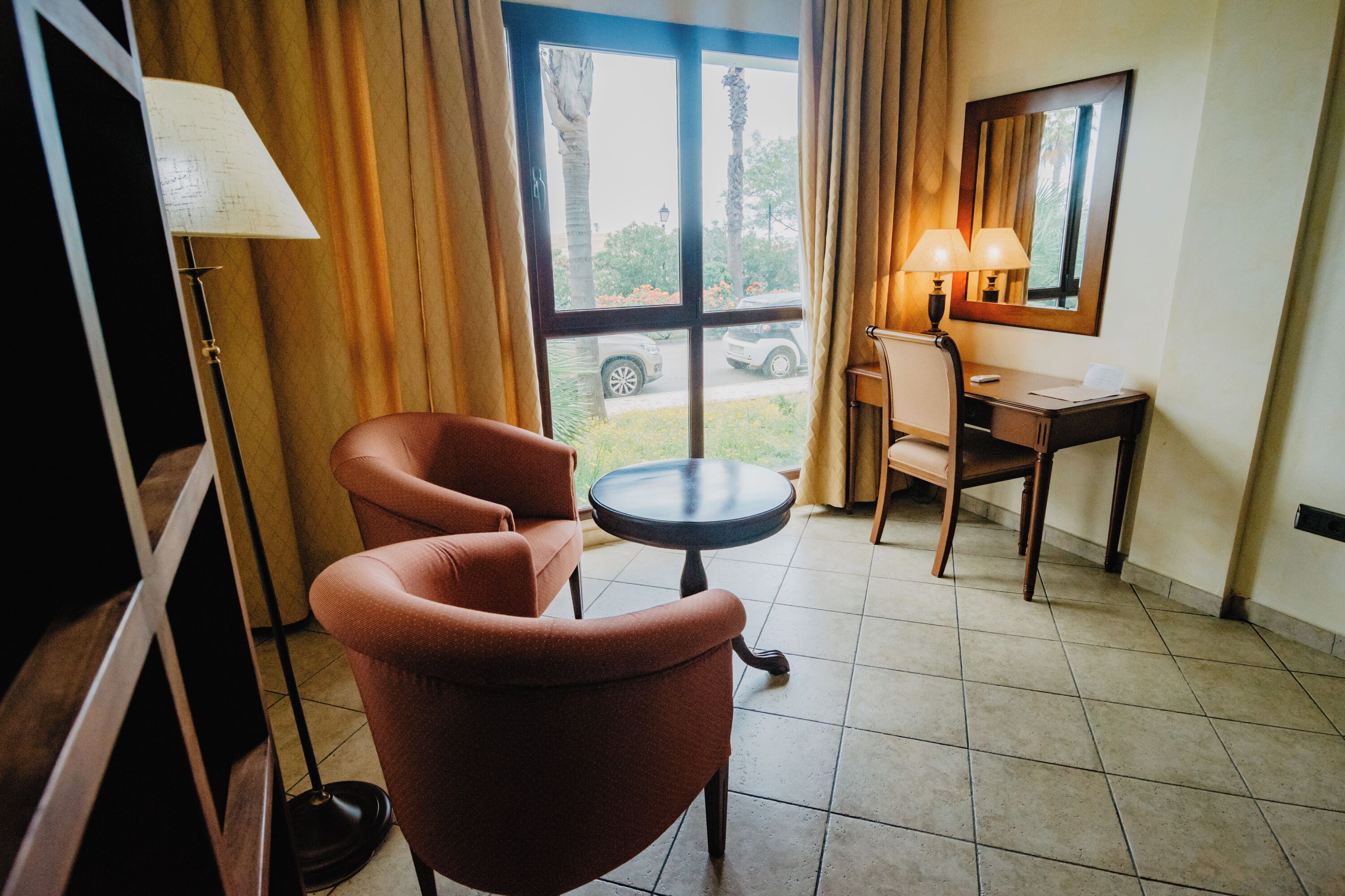 A hotel room with a red armchair, a small round table, a desk with a lamp, and a window with a view of the outside.