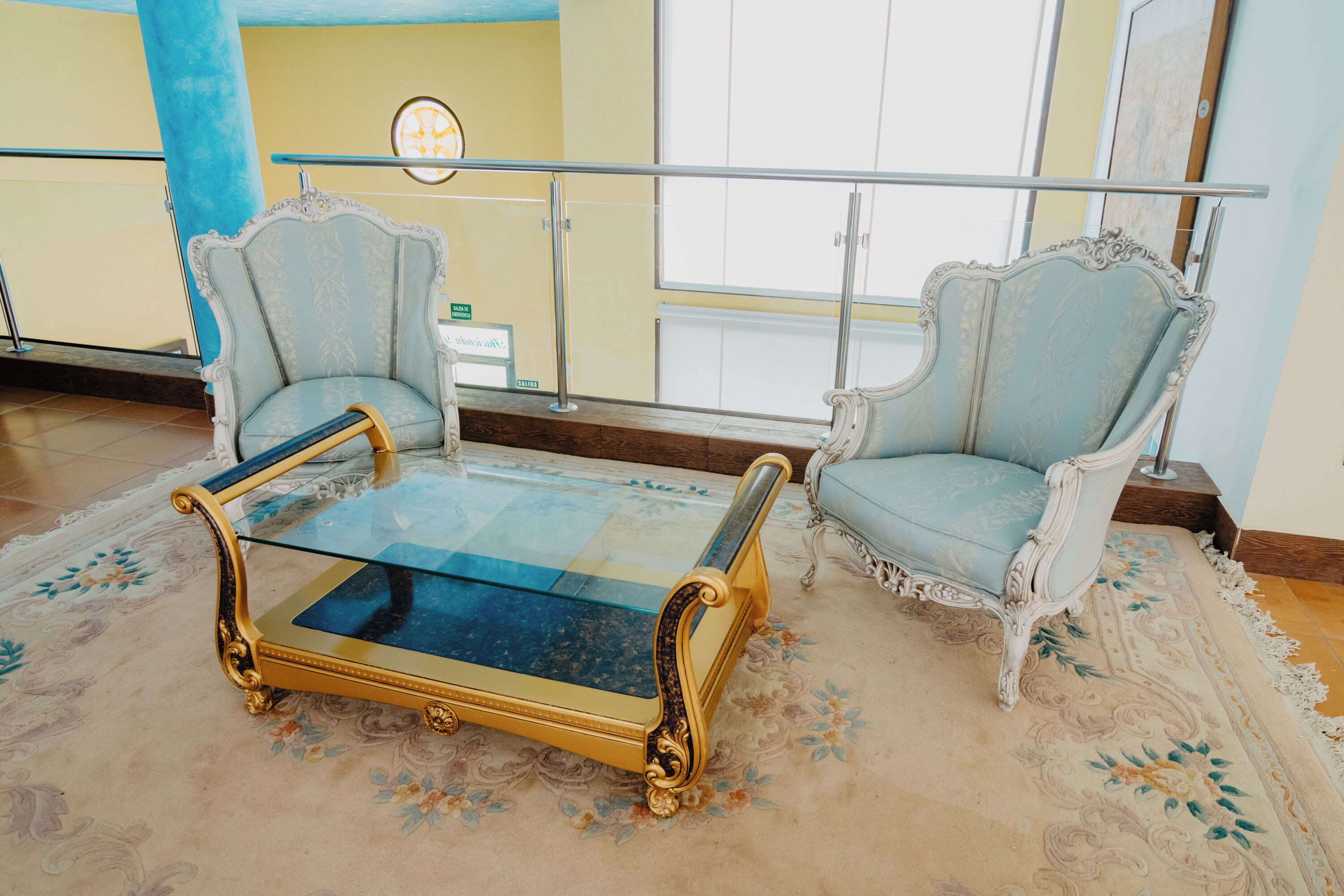 Una habitación con dos sillones ornamentados, una mesa de centro con superficie de vidrio y una alfombra de estampado floral.