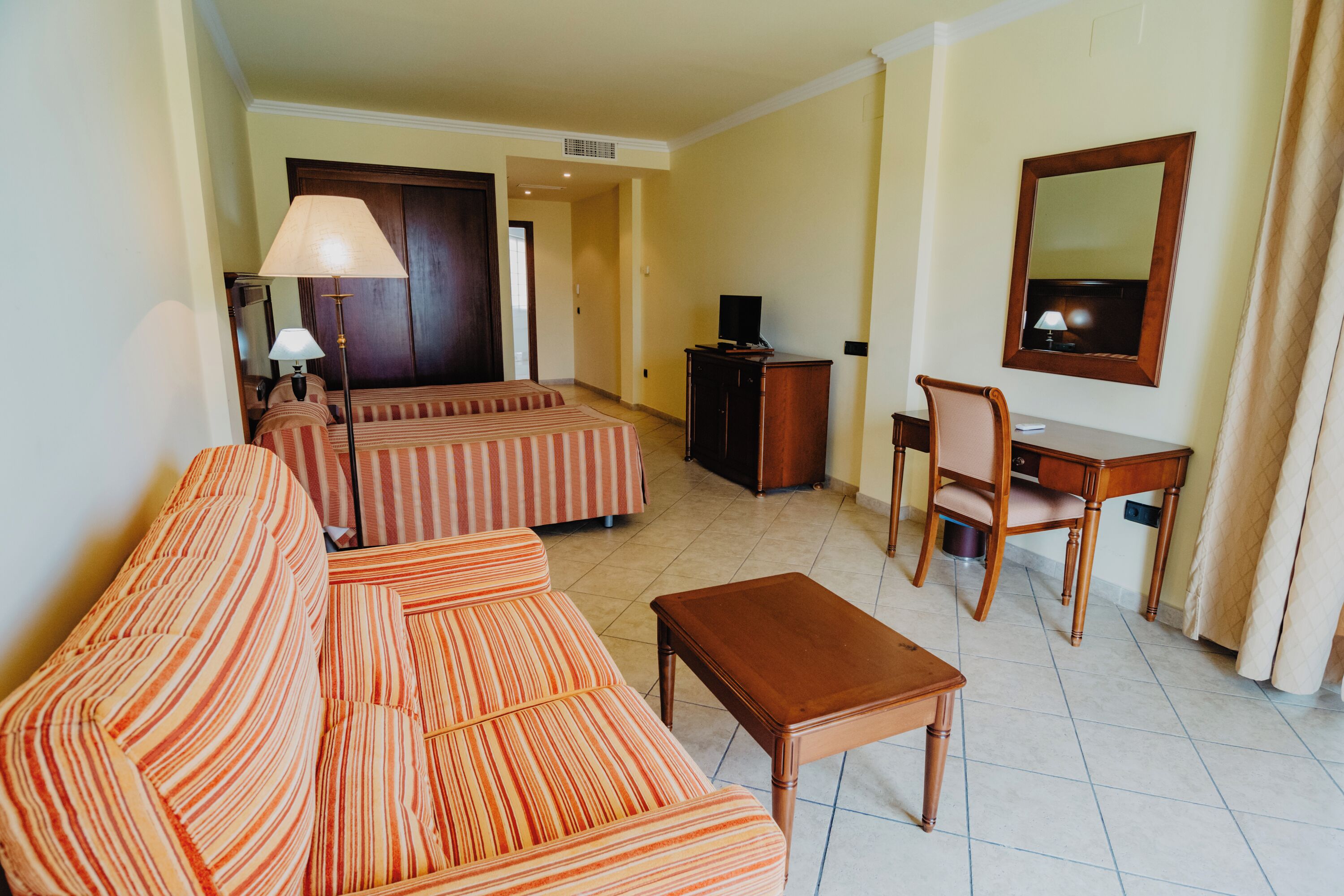 A hotel room with a striped sofa set, a wooden coffee table, a desk with a chair, a television, and a mirror on the wall.