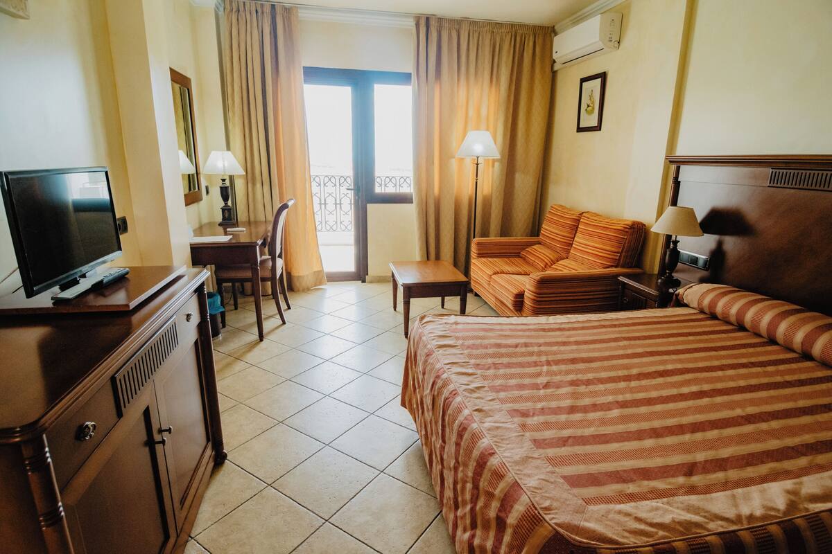 A hotel room with a bed, a desk with a television, a chair, a sofa, and a balcony with curtains.