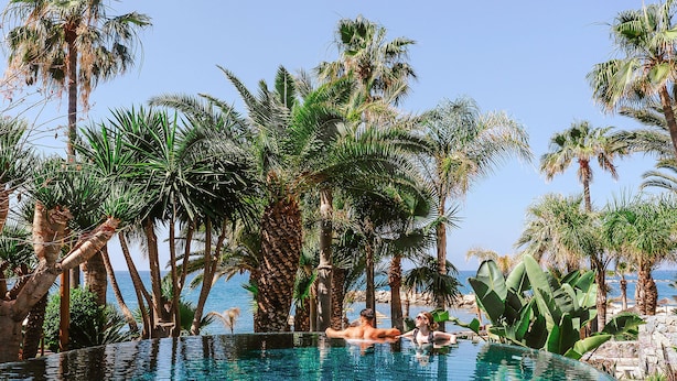 A clear blue pool surrounded by palm trees and a view of the sea. - Amathus Residences (Limassol)