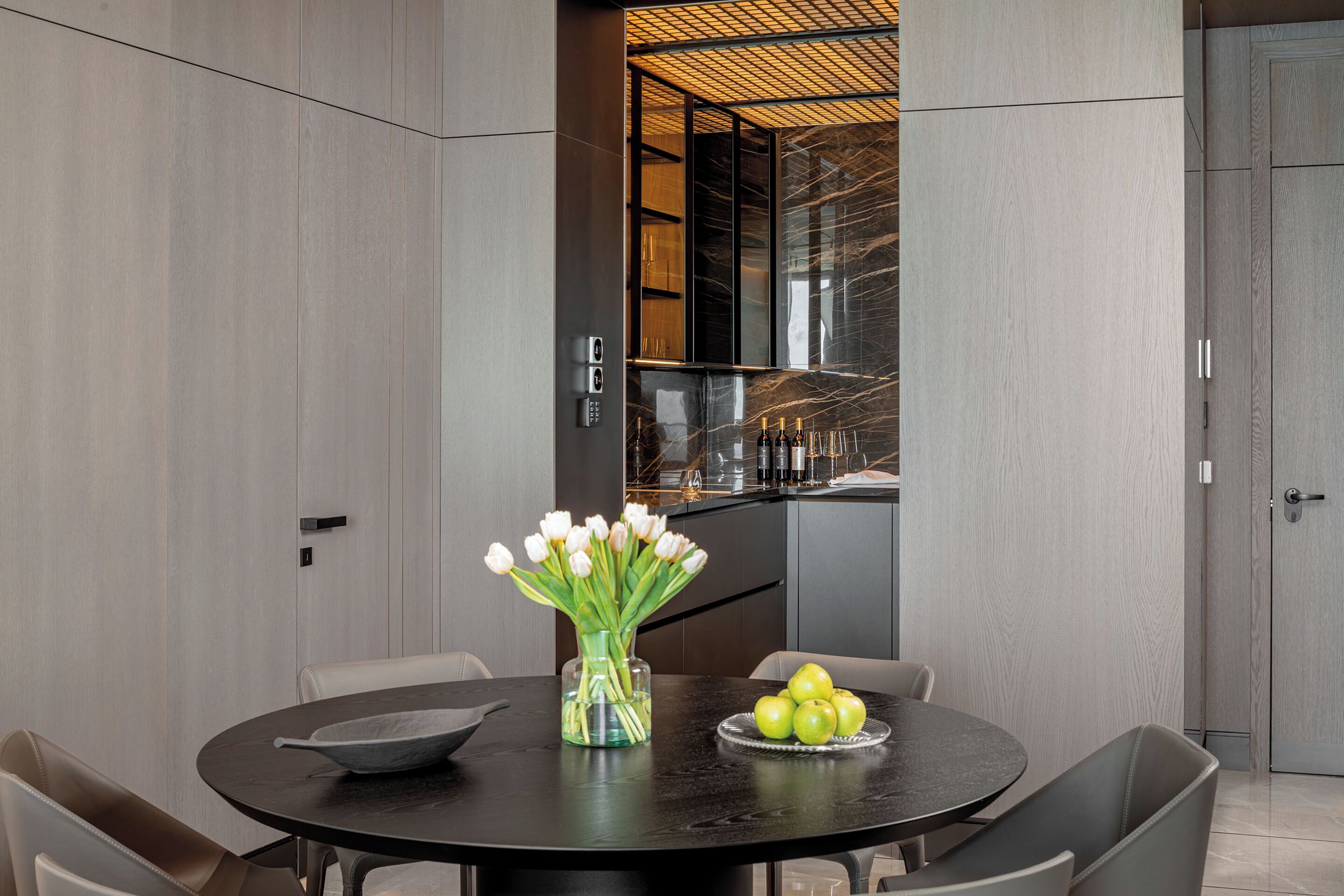 A modern dining area with a round table, chairs, a vase with flowers, and a bowl of apples.