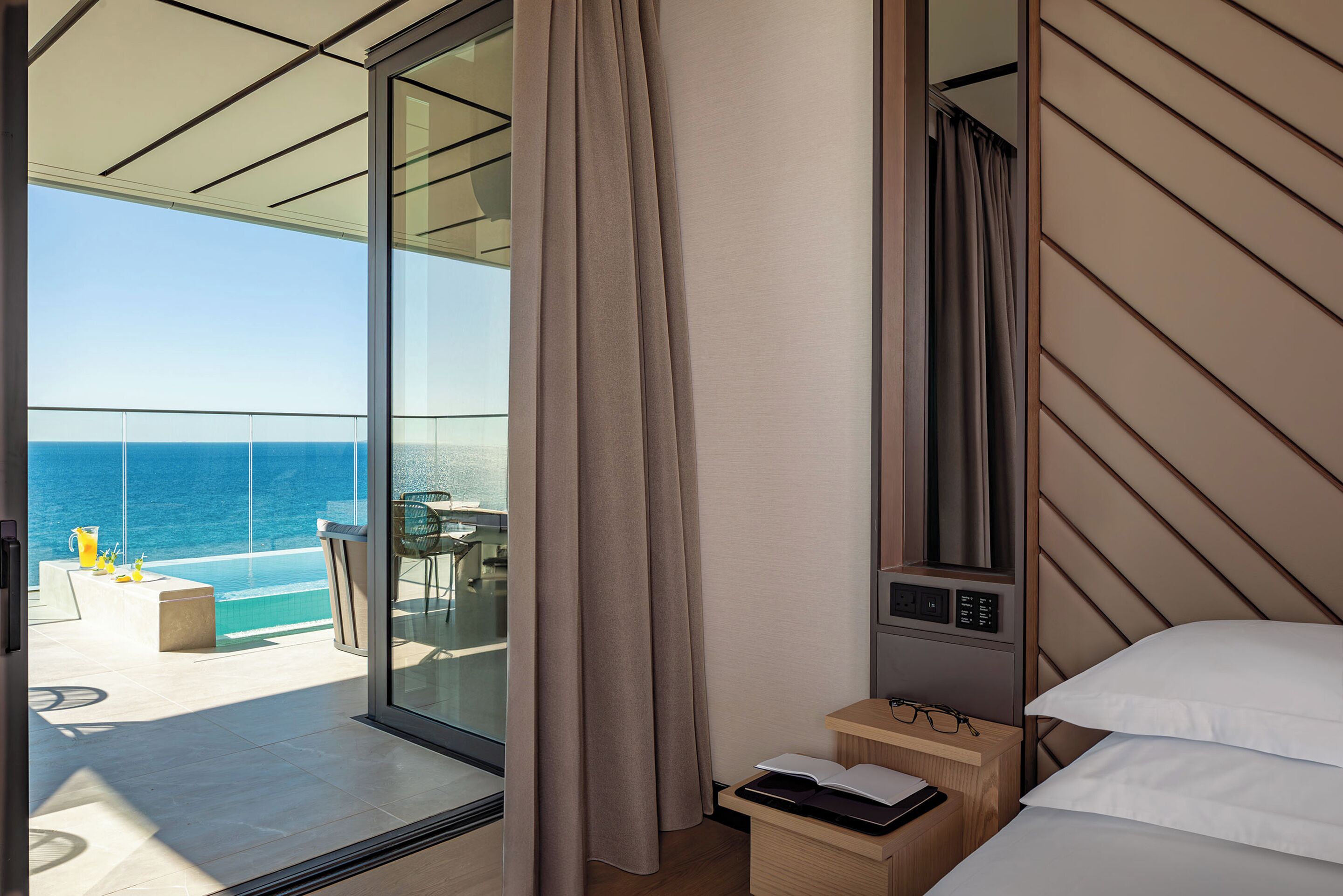 A modern hotel room with a large sliding glass door leading to a balcony with a pool and ocean view.