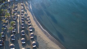An aerial view of a beach with rows of sun loungers and umbrellas, a clear blue sea, and a rocky shoreline. - Amathus Residences (Limassol)