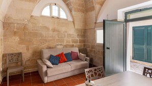 A room with a stone wall, a beige sofa with colorful cushions, a wooden table, and a door with green shutters. - 3936 Corte dei Falconi by Barbarhouse (Lecce)