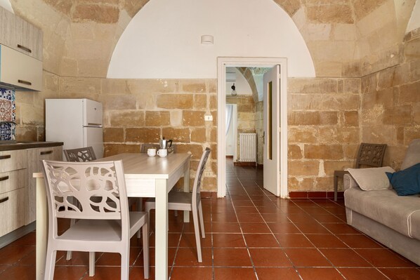 A kitchen with stone walls, a dining table with chairs, a white refrigerator, and a sofa with a cushion. - 3936 Corte dei Falconi by Barbarhouse (Lecce)