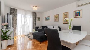 A modern living room with a black sofa, white dining table, and a glass coffee table. There are framed artworks on the wall and a potted plant on the floor.