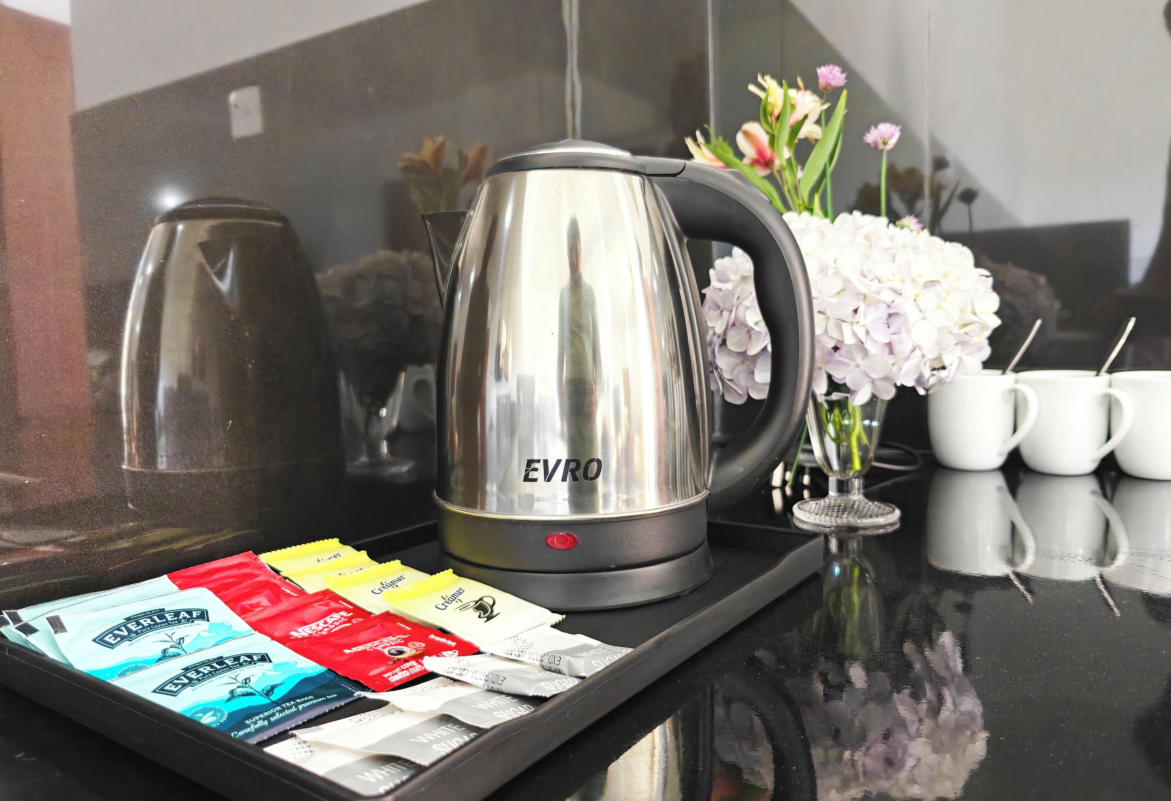 A stainless steel EVRO kettle on a tray with coffee packets, a vase with flowers, and white mugs on a reflective surface.