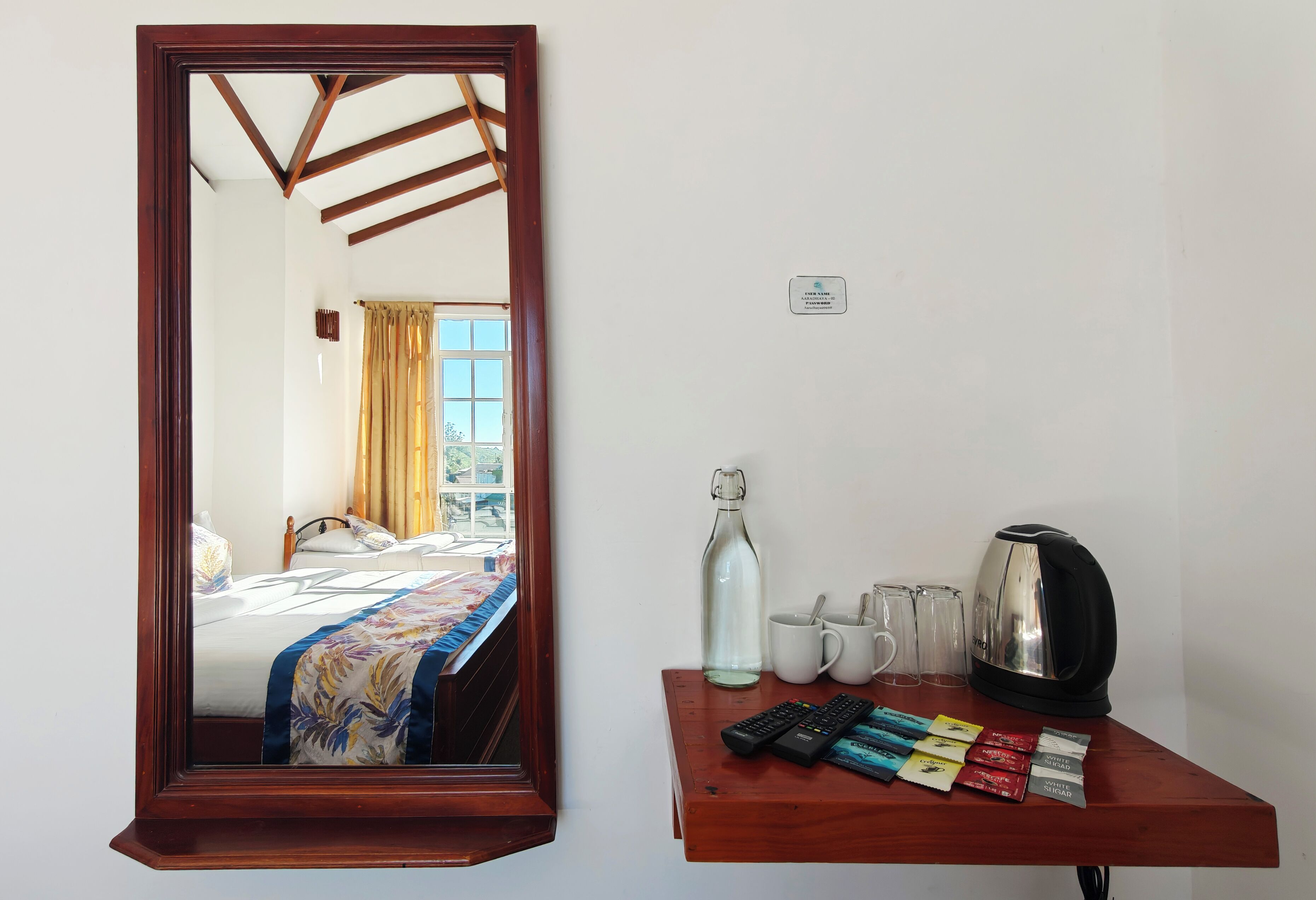 A hotel room with a wooden-framed mirror, a bed with a floral bedspread, a wooden side table with a kettle, mugs, and remote controls.