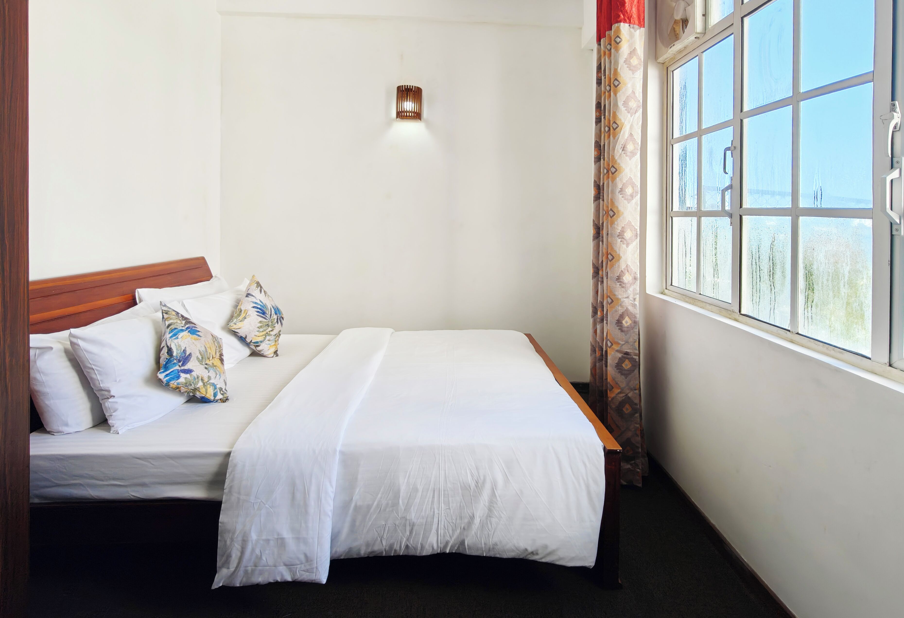 A single bed with white bedding, a wooden headboard, a wall-mounted light, and a window with patterned curtains.