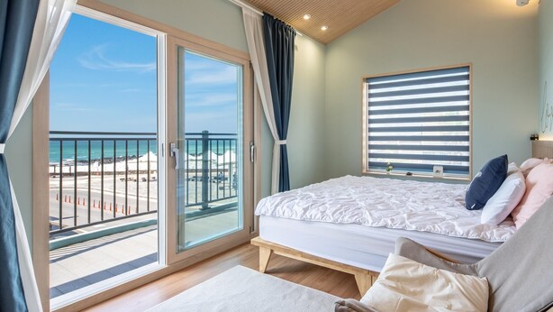 Un dormitorio con un gran ventanal corredera que da a un balcón, una cama con ropa blanca y vistas a la playa.