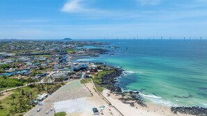 Vista aérea de un pueblo costero con playa, zona de estacionamiento y aerogeneradores a lo lejos.