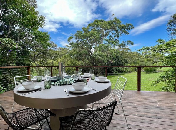 A round table set for dining on an outdoor deck with chairs, surrounded by lush greenery and trees.