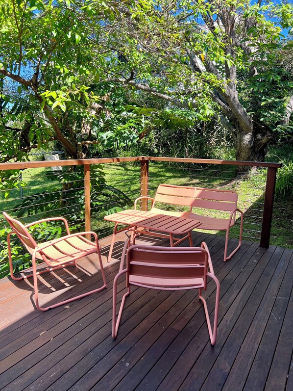 Eine Holzterrasse mit Metallstühlen und -tischen, umgeben von üppigem Grün und Bäumen.