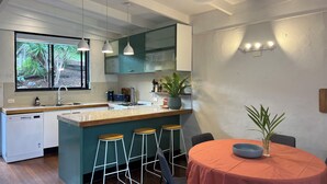 A modern kitchen with a central island, bar stools, a dining table with a red tablecloth, and a view of greenery outside. - Surfers shack - peaceful & minutes walk to the beach in stunning Nambucca (Nambucca Heads)