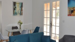 A modern living room with a blue sofa, a round coffee table, and a glass dining table with chairs. There is a door with glass panels and a poster on the wall. - Appartement 3 Chambres + Terrasse de 60 m2 Au1er Étage D'une Villa à Golfe Juan (Vallauris)