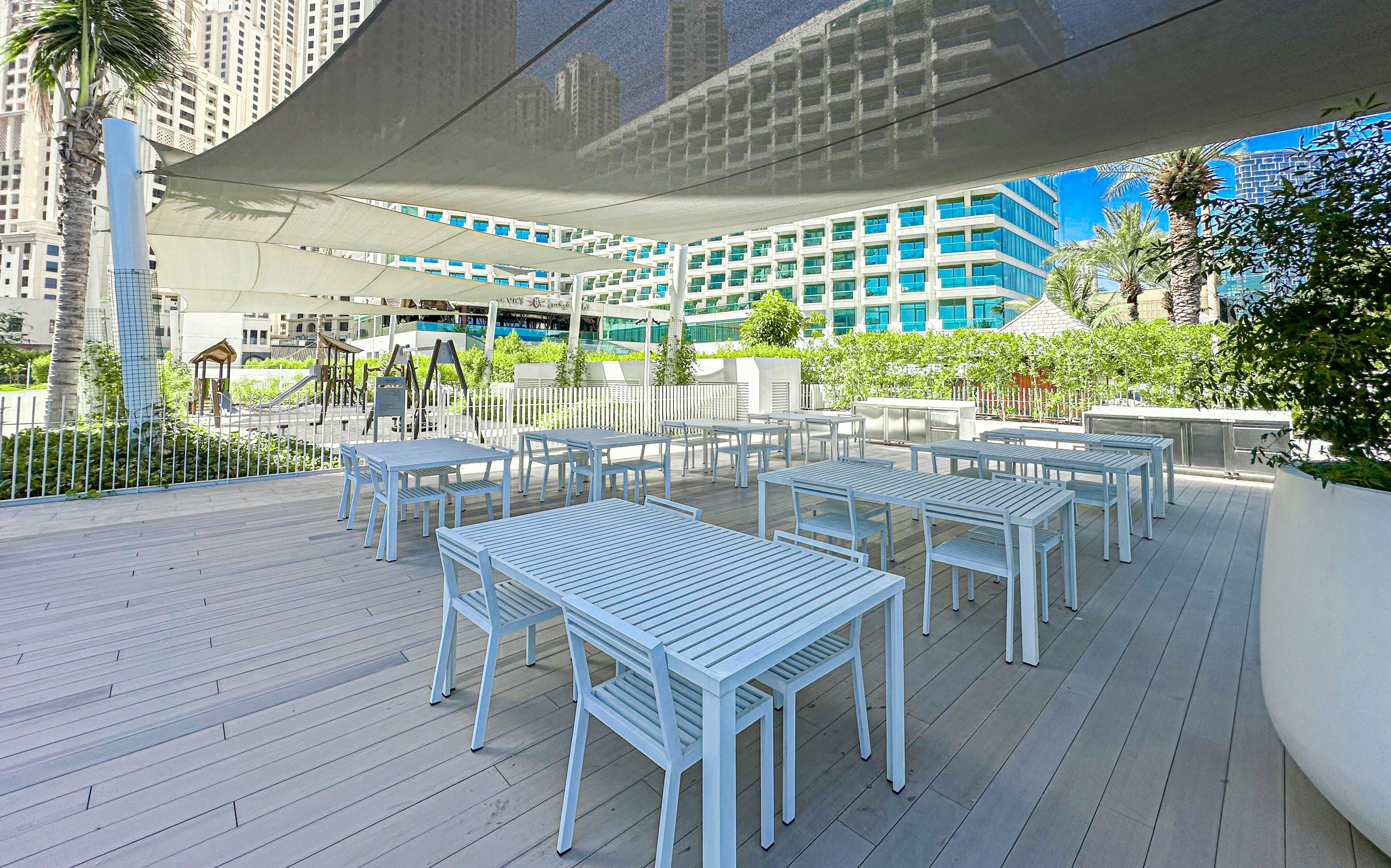 Une terrasse extérieure avec des tables et des chaises blanches, entourée de verdure et avec un bâtiment moderne en arrière-plan.