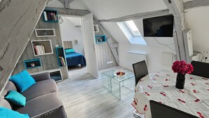 Une pièce de style loft avec un plafond en pente, une lucarne, un lit avec une literie bleue, un canapé avec des coussins bleus, une table à manger recouverte d’une nappe à motifs floraux et un téléviseur fixé au mur.