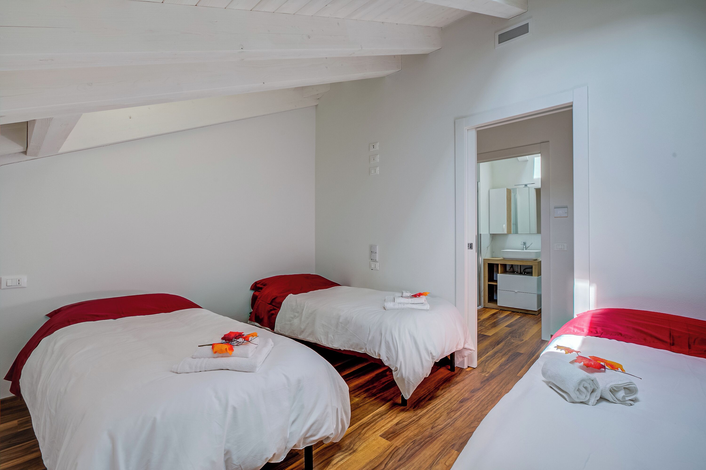 A hotel room with three beds, each with a red headboard and white bedding. There is a wooden floor and a white ceiling with exposed beams. A bathroom is visible in the background.