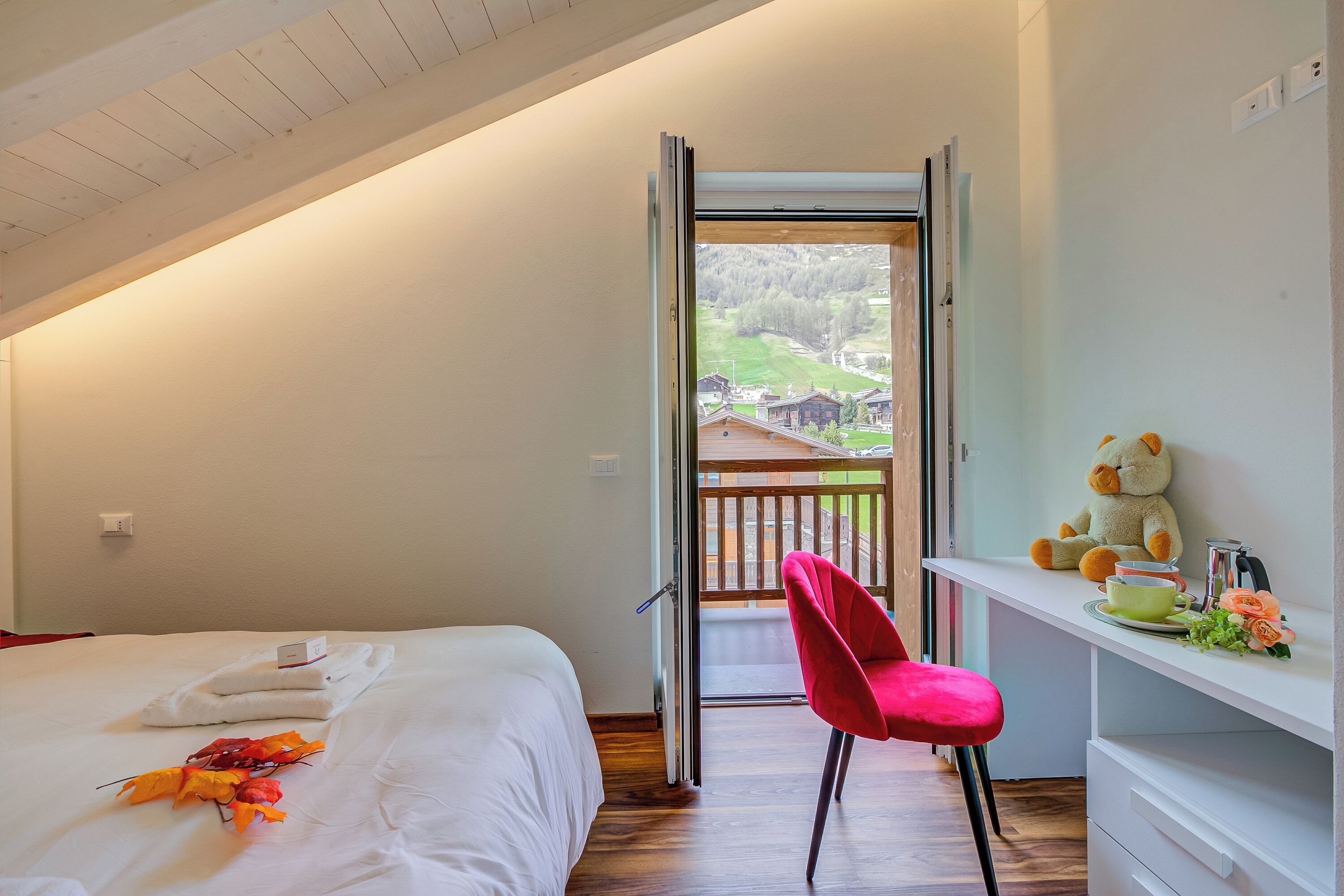 Un dormitorio con cama, escritorio, silla, un oso de peluche y vistas a un balcón.