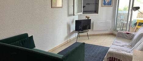A room with a green sofa, a bed with white bedding, a TV on a stand, and a window with a view of the outside.