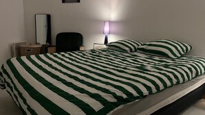 Een bed met groen-wit gestreepte lakens, een bedlamp, een stoel, een klein tafeltje en een aan de muur hangend schilderij.