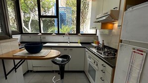 Una cocina pequeña con ventana, estufa, pileta y un taburete.
