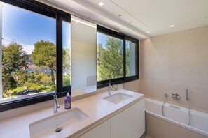 A modern bathroom with two sinks, a bathtub, and a large window offering a view of greenery and a distant town.