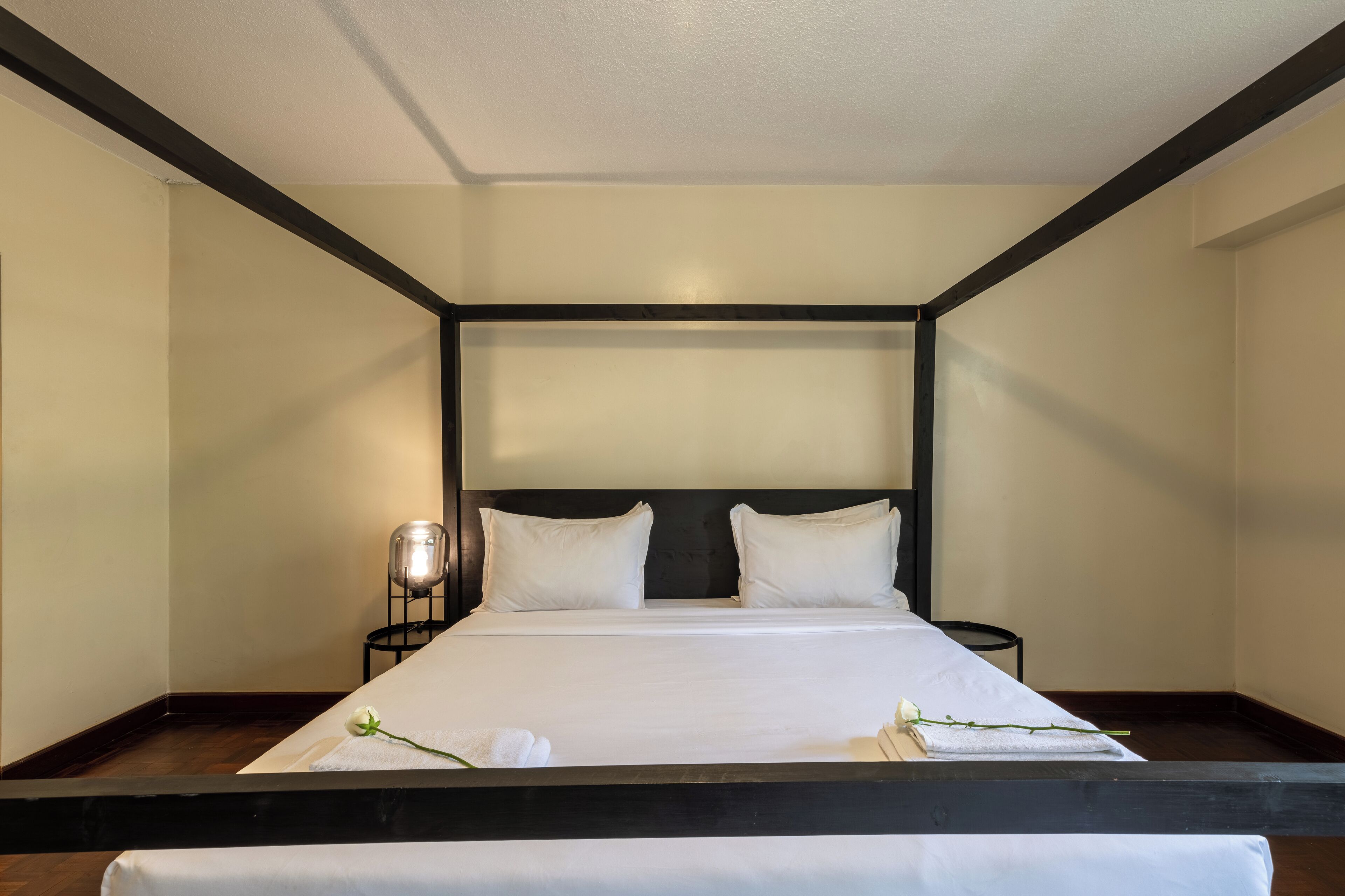 Una cama bien tendida con sábanas y almohadas blancas, una mesita de noche con una lámpara, y un pequeño florero con una flor blanca.