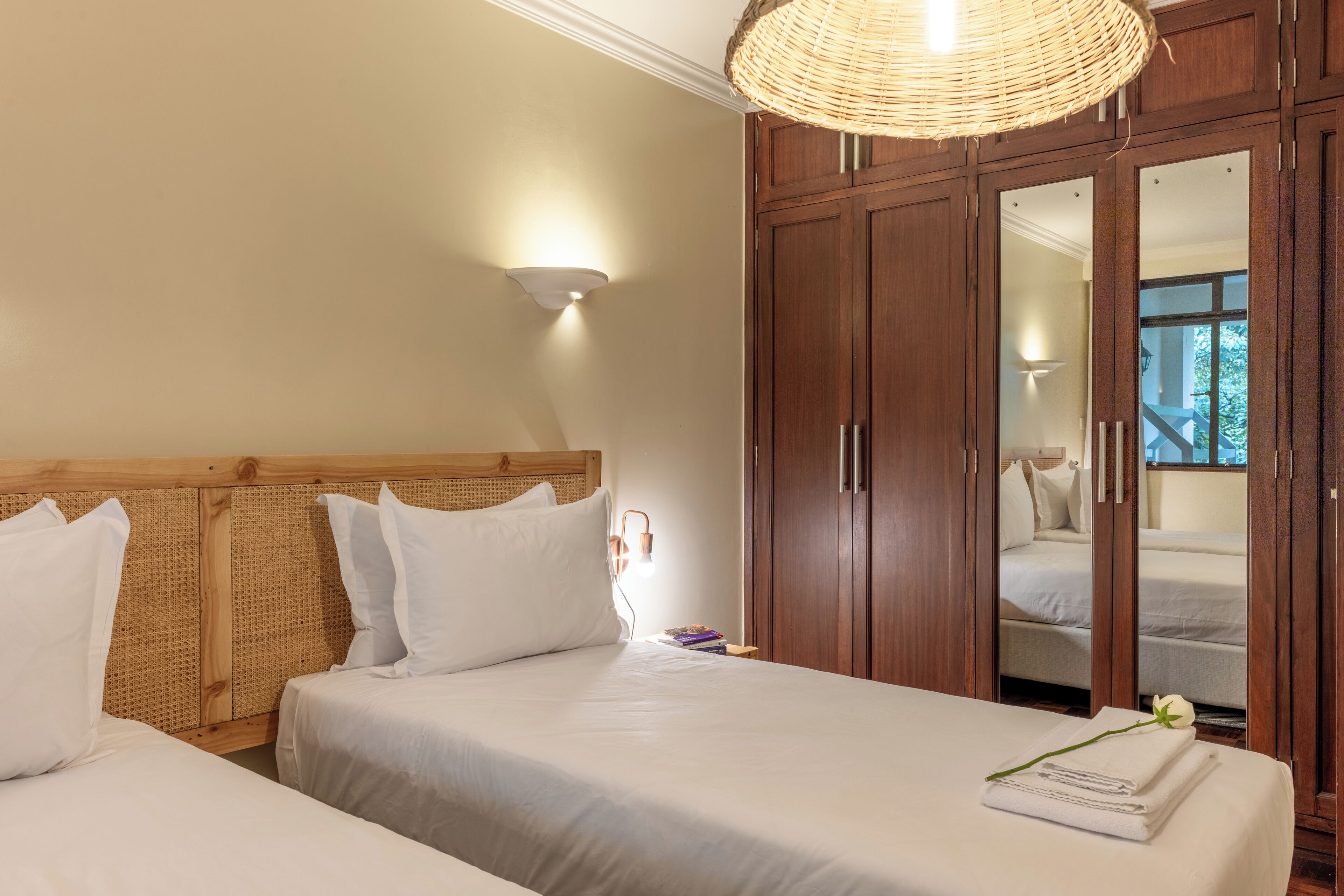 A hotel room with two beds, a wooden wardrobe, a woven ceiling light fixture, and a wall-mounted light.