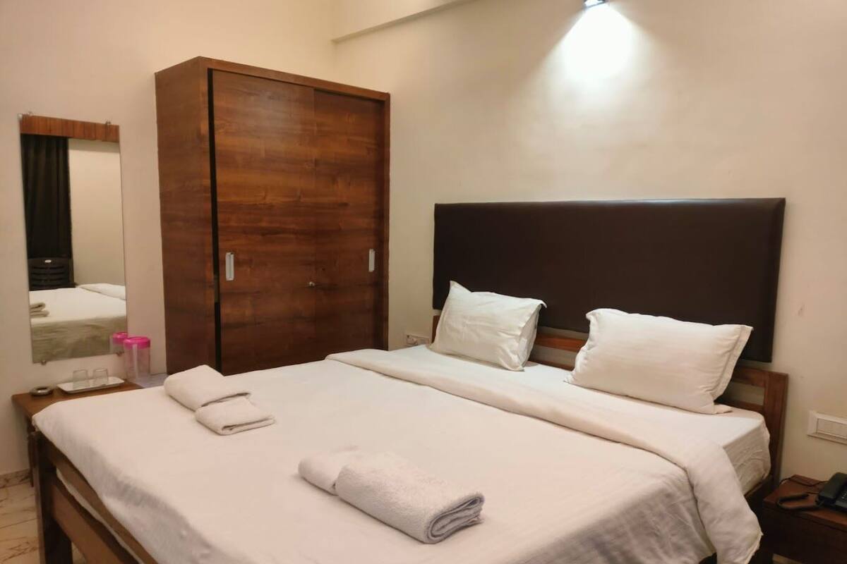 Una cama bien hecha con sábanas blancas y dos almohadas, un armario de madera, una mesita de noche con lámpara y un espejo en la pared.