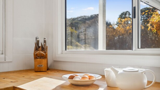 Una encimera de cocina con una tabla de cortar de madera, un bol de huevos, una tetera blanca y un bloque de cuchillos.
