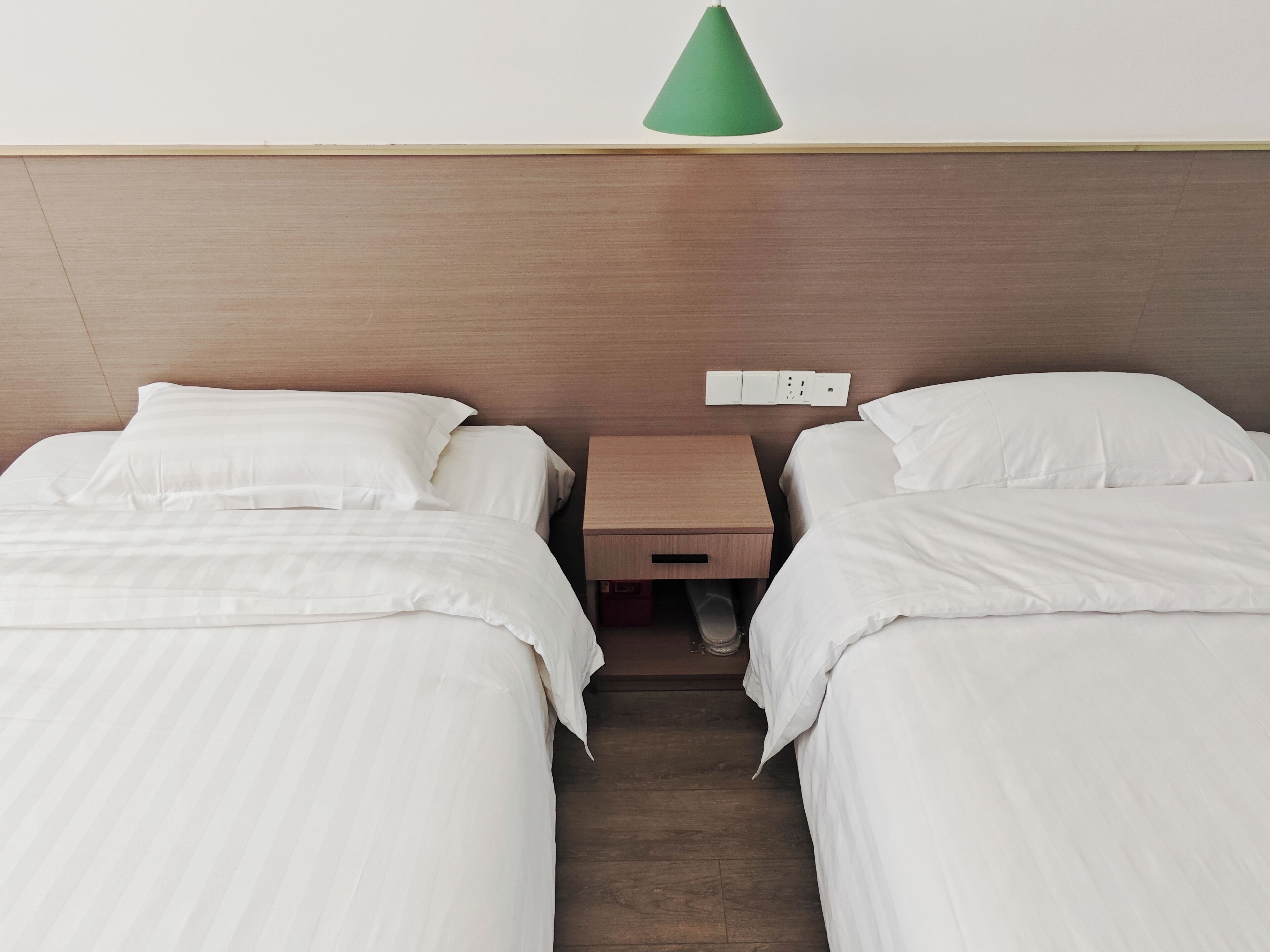 Quarto de hotel com duas camas, uma cabeceira de madeira, uma mesinha de cabeceira e um candeeiro suspenso verde.