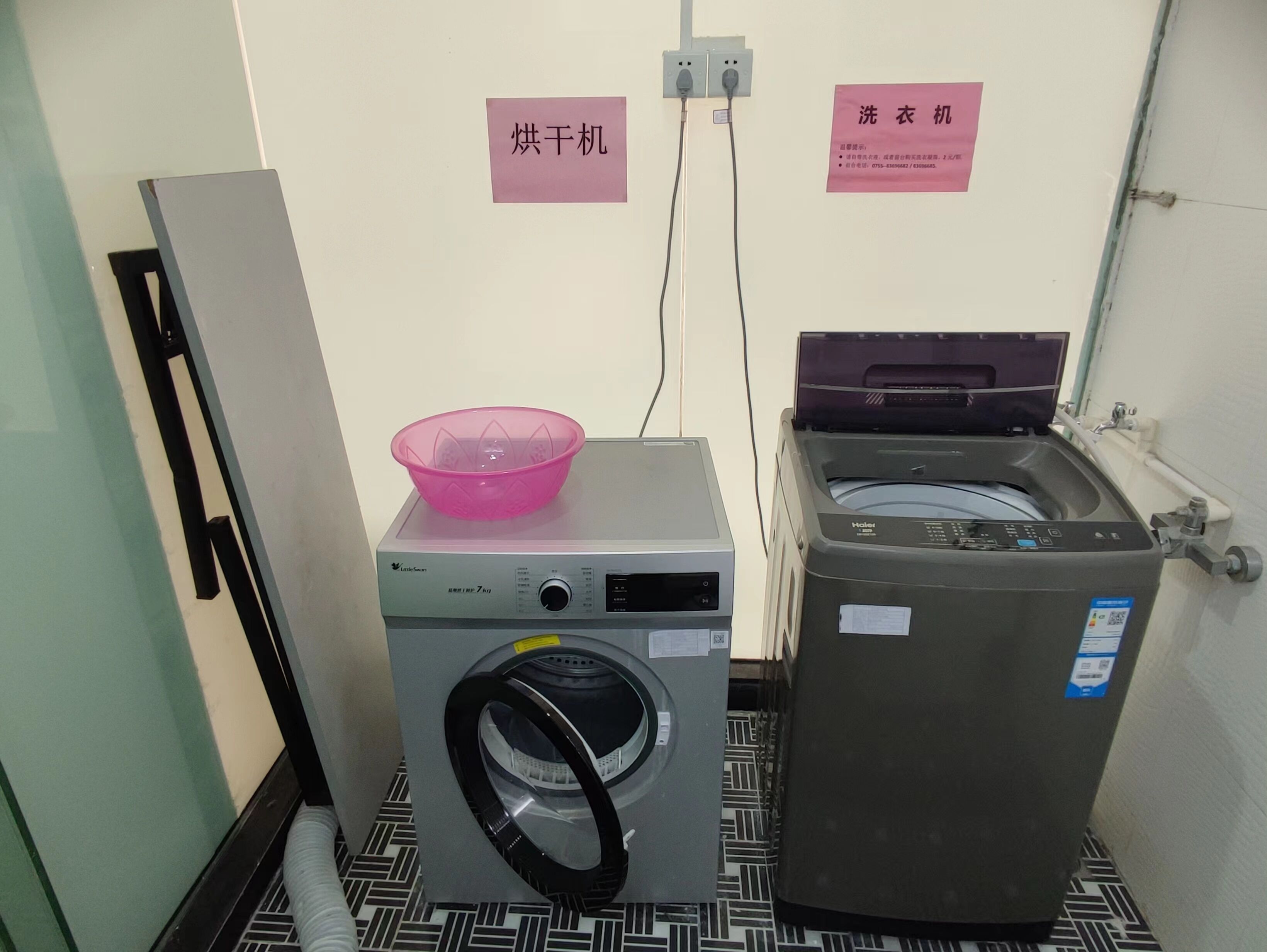 A laundry room with a front-loading washing machine and a top-loading dryer. There is a pink laundry basket on the washing machine, and a sign on the wall indicating the location.